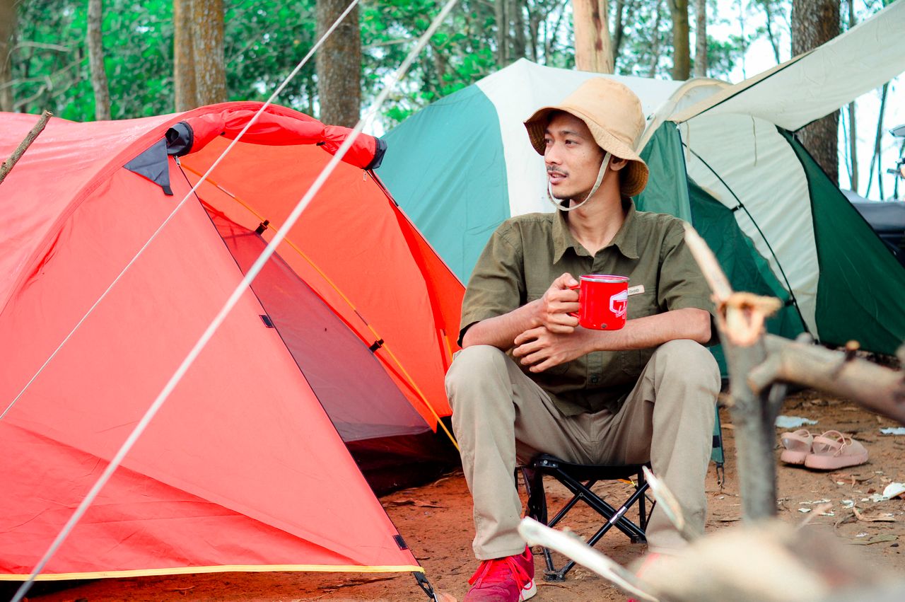Man sitting outdoors with a red cup near tents in the forest, enjoying camping.