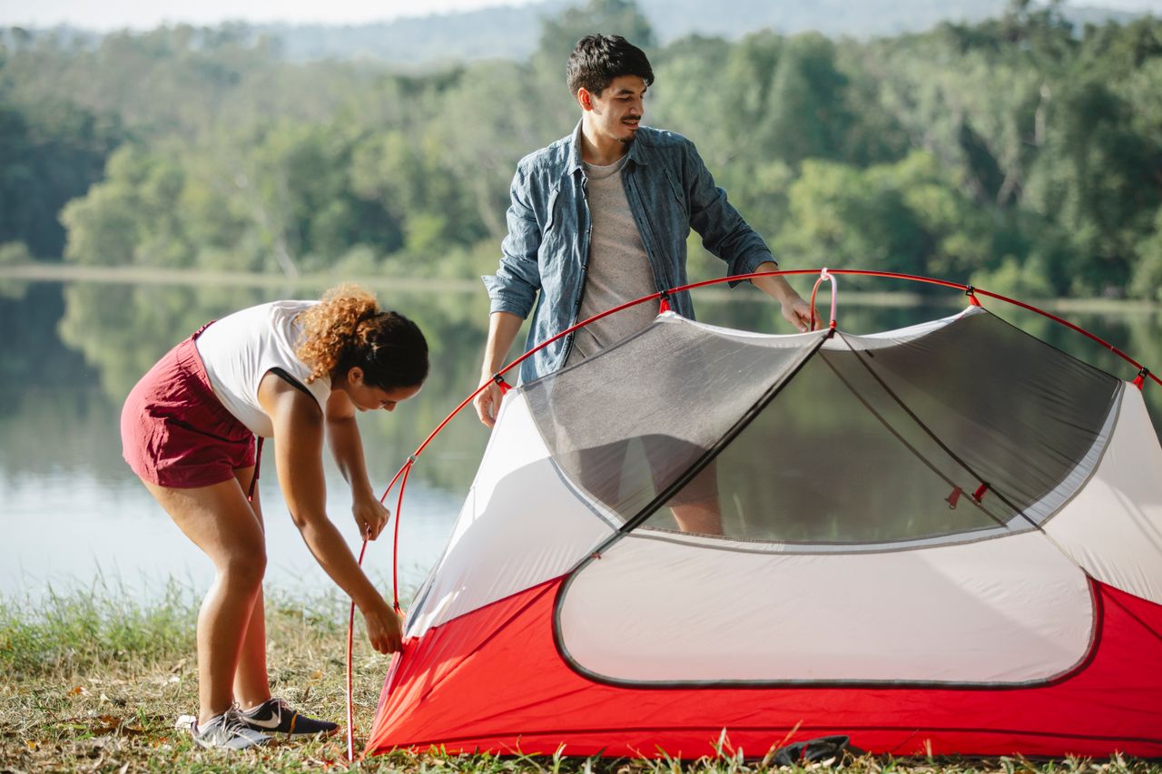 Full body young diverse couple of travelers putting up colorful tent on grassy river bank
